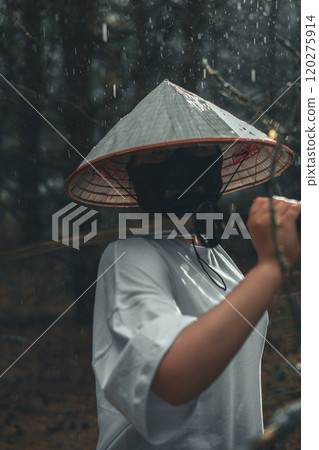 A mysterious man in a black mask and a conical hat holds a katana in a rainy forest. The samurai exudes calm and strength as he holds the katana loosely on his shoulder. 120275914