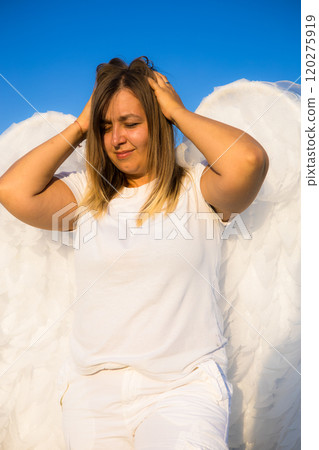An angel in despair holds her head from what is happening in the world. A blonde in white clothes and with big white angel wings touches her hair with both hands under a clear blue sky. 120275919