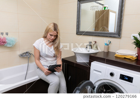 A woman relaxes in her bathroom with a tablet, creating a harmonious space by a washing machine A woman relaxes in her bathroom with a tablet, creating a harmonious space by a washing machine 120279166