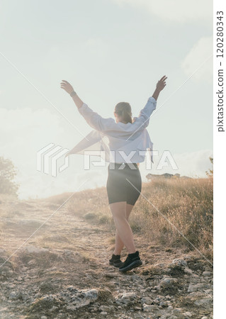 Woman, Field, Sunset - Woman in a white shirt and black skirt dances on a hill at sunset with arms raised. 120280343