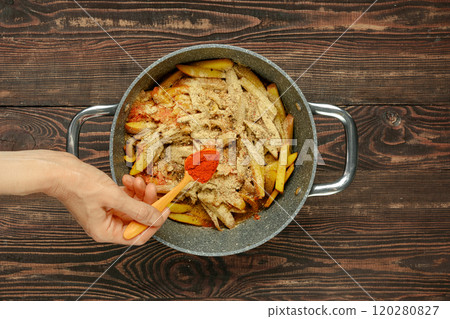 Hand holds a wooden spoon, adding smoked paprika to a pot filled with bell peppers and potatoes Hand holds a wooden spoon, adding smoked paprika to a pot filled with bell peppers and potatoes 120280827