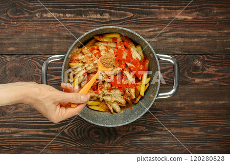 A hand adds coriander to a pot filled with sliced potatoes and bell peppers 120280828