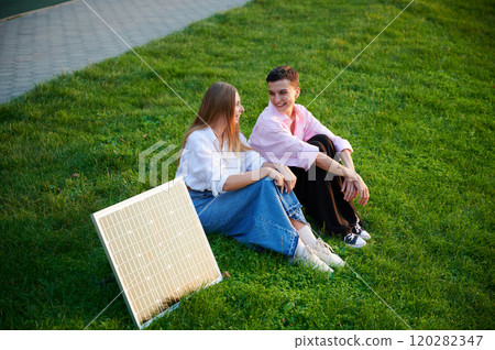 Young women using solar panel for charging, sit on grassy lawn, smiling and talking, with solar panel placed nearby. Girls dressed casually, enjoying sunny, relaxed moment outdoors. Young women using solar panel for charging, sit on grassy lawn, smiling and talking, with solar panel placed nearby. Girls dressed casually, enjoying sunny, relaxed moment outdoors. 120282347