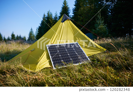 Yellow tent with solar panel set up in grassy field surrounded by dense evergreen trees under clear blue sky. Eco-friendly camping, integration of renewable energy in outdoor activities. Yellow tent with solar panel set up in grassy field surrounded by dense evergreen trees under clear blue sky. Eco-friendly camping, integration of renewable energy in outdoor activities. 120282348