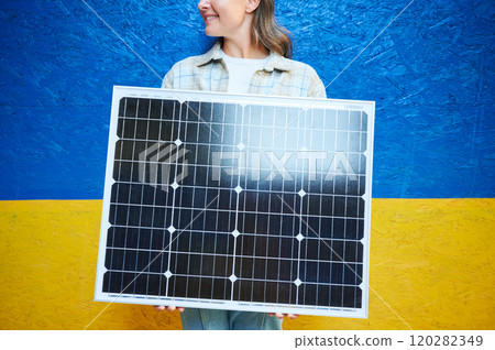 Woman holding solar panel for charging. Smiling female wears plaid shirt over white top, standing against vibrant blue and yellow Ukrainian flag background, symbolizing sense of pride and energy. Woman holding solar panel for charging. Smiling female wears plaid shirt over white top, standing against vibrant blue and yellow Ukrainian flag background, symbolizing sense of pride and energy. 120282349