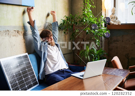 Bearded man stretches with arms overhead while working on laptop at wooden table. Solar panel nearby, emphasizing eco-friendly workspace that combines technology and sustainability in cozy environment 120282352