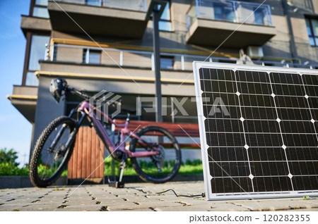 Pink electric mountain bike parked next to wooden bench, connected to solar panel for charging on a blurred background. Bike rests on paved area surrounded by greenery and grass. 120282355