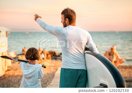 Father and daughter are walking along the beach, looking for a place to swim and surf in the sea. Back view. Sunset light. Summer vacation 120282530