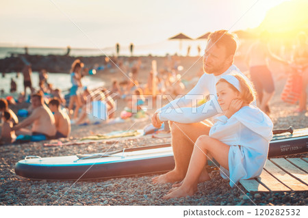 Father and him daughter sits talking and relax at the pebble beach. Sup board lie near man. Sunset light. Family vacation at the sea. Summertime 120282532