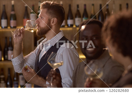 Side view of Caucasian professional sommelier tasting chilled white wine while his highly skilled colleagues communicating happily 120282680