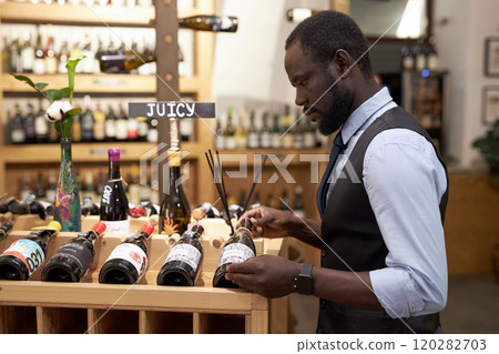Side view of African American sommelier looking at wine while putting it on wooden rack 120282703