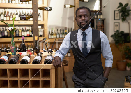 Medium close up portrait of African American sommelier in smart clothes standing next to wooden rack with wine bottles 120282705