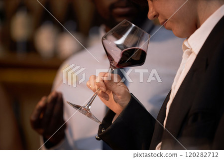 Medium close up of unrecognizable woman trying to scent red wine while unrecognizable African American sommelier telling about notes and body in blurred background Medium close up of unrecognizable woman trying to scent red wine while unrecognizable African American sommelier telling about notes and body in blurred background 120282712