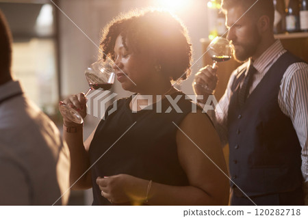 Mixed race mature woman inhaling scent of red wine with closed eyes during tasting in wine cellar, she illuminated with sunrays 120282718