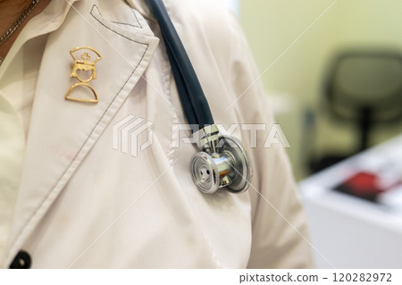 Close-up of a doctor's white coat with a stethoscope hanging around the neck. The background is softly blurred, showing a clinical or office setting with light green hues. Close-up of a doctor's white coat with a stethoscope hanging around the neck. The background is softly blurred, showing a clinical or office setting with light green hues. 120282972