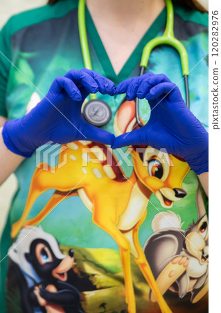 A healthcare professional wearing blue gloves forms a heart shape with their hands over a shirt featuring a colorful cartoon character, with a green stethoscope draped around their neck. A healthcare professional wearing blue gloves forms a heart shape with their hands over a shirt featuring a colorful cartoon character, with a green stethoscope draped around their neck. 120282976