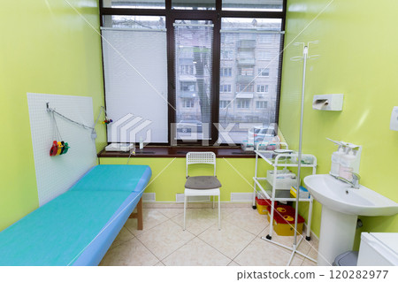 A bright medical examination room with a green accent wall, blue patient bed, window with blinds, chair, medical supplies on a cart, IV stand, and sink, creating a clean and organized environment. A bright medical examination room with a green accent wall, blue patient bed, window with blinds, chair, medical supplies on a cart, IV stand, and sink, creating a clean and organized environment. 120282977