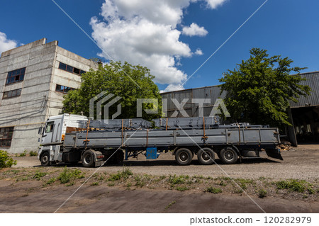 A large flatbed truck loaded with metal beams is parked near industrial buildings on a sunny day. Trees and blue sky with clouds are visible in the background. The area has a rugged, industrial vibe. 120282979