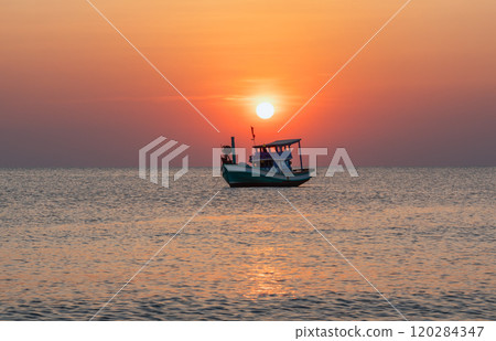 A fishing boat on the water under the setting sun, reflected in the calm ocean waves. The warm sunset colors add romance and tranquility to the scene A fishing boat on the water under the setting sun, reflected in the calm ocean waves. The warm sunset colors add romance and tranquility to the scene 120284347