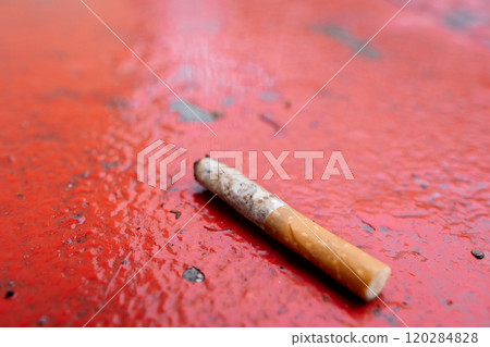 Close-up of a discarded cigarette lying on a wet red ground surface with visible water droplets Close-up of a discarded cigarette lying on a wet red ground surface with visible water droplets 120284828