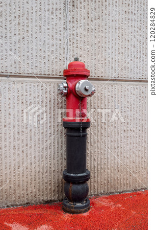 Red fire hydrant with black base mounted on a textured concrete wall with red ground surface 120284829