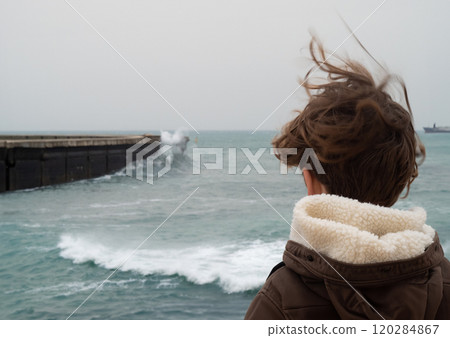 Caucasian boy with brown hair wearing a jacket filmed from the back watching stormy waves crash against a pier on a cloudy day 120284867