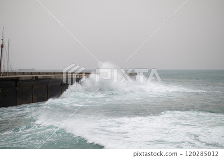 Rough ocean waves crashing powerfully against a concrete pier on a foggy day, with water spraying over the edge and creating a dramatic scene 120285012