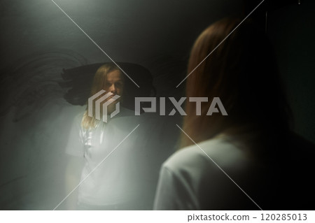 Woman with straight blonde hair staring at her reflection in a foggy mirror, creating a somber and introspective mood 120285013