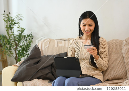 Satisfied young asian woman making a purchase online with credit card and digital tablet at home 120288011
