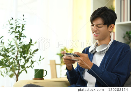 Excited young Asian man with headphones playing video game on smartphone Excited young Asian man with headphones playing video game on smartphone 120288154