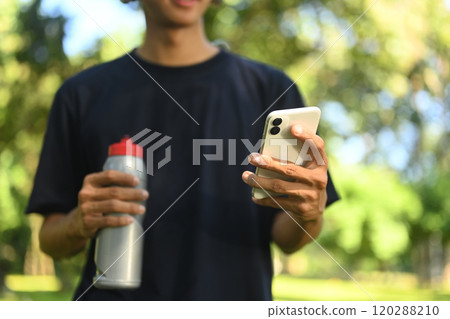 Young athlete holding water bottle and checking his smartphone in the park Young athlete holding water bottle and checking his smartphone in the park 120288210
