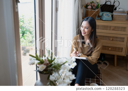 Beautiful young woman writing in notebook while looking out the window Beautiful young woman writing in notebook while looking out the window 120288323