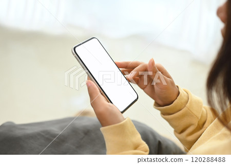 Woman in yellow sweater holding smartphone with white empty display sitting on sofa 120288488