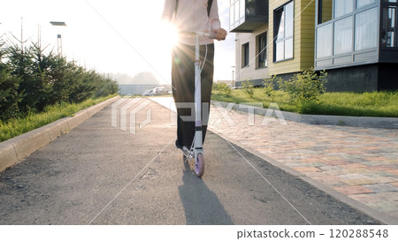 Cheerful girl with a backpack rides her scooter through a quiet residential street on her way to school in a modern neighborhood Cheerful girl with a backpack rides her scooter through a quiet residential street on her way to school in a modern neighborhood 120288548