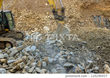 Large excavator digs into rocky area, lifting heavy boulders debris at construction site, with dust dirt being displaced during operation. 120290457