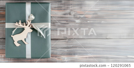A Christmas gift wrapped in green paper with a white ribbon and a wooden reindeer ornament, placed on a wooden table, viewed from above 120291065