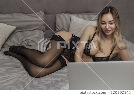 Sexy woman in black lingerie lies on the bed and uses a laptop. Sex model concept on webcam and dating site 120292143