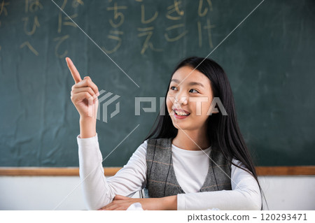 A young girl is sitting at a desk in a classroom with a green chalkboard behind her. She is pointing to the board with her finger, possibly indicating something important or interesting 120293471
