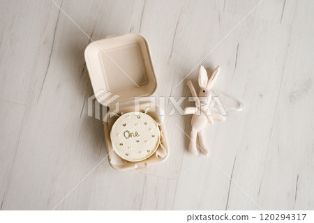 Toy rabbit on white surface next to a container with a cake inside, labeled with the number one Toy rabbit on white surface next to a container with a cake inside, labeled with the number one 120294317