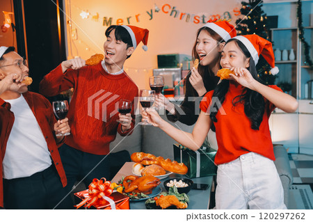 Group of young Asian man and women as friends having fun at a New Year's celebration, holding gift boxes standing by Christmas tree decoration, midnight countdown Party at home with holiday season. 120297262