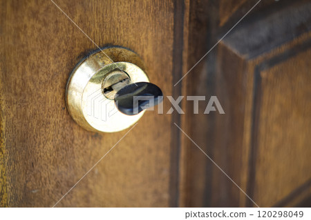 Close-up of a worn key inserted into an old, yellowed wooden door, highlighting a vintage keyhole and weathered knob. The rustic textures and faded colors emphasize age 120298049