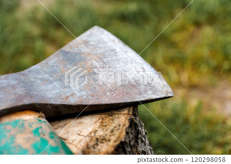 Close-up of an old, weathered hatchet embedded in a tree stump, with a blurred green background highlighting the rugged details of the axe. The aged metal and worn handle convey themes of rustic work Close-up of an old, weathered hatchet embedded in a tree stump, with a blurred green background highlighting the rugged details of the axe. The aged metal and worn handle convey themes of rustic work 120298058