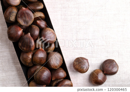 Sweet chestnuts in cardboard punnet on linen, from above, with empty space for text. Castanea sativa, raw, whole and unshelled. Can be eaten raw, candied, cooked, or roasted as warm snack on streets. 120300465