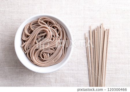 Chilled, boiled soba noodles in a white bowl on linen fabric. Thin Japanese noodles, made primarily from buckwheat flour, with wheat flour mixed in. With a bunch of dried noodles on the right side. 120300466