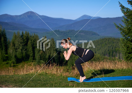 Woman practicing yoga outdoors in the mountains in a serene, natural setting. Female performing yoga pose on blue mat, with backdrop of beautiful mountain landscape at sunrise or sunset. 120307966