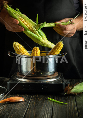 Hands skillfully shuck fresh corn, revealing tender yellow kernels. The pot simmers on the stove, surrounded by earthy ingredients, evoking a warm cooking atmosphere 120308367