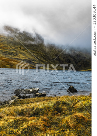 A calm lake reflects misty hills and rocky formations, surrounded by lush grassland. This picturesque setting captures the beauty of nature in a remote region. A calm lake reflects misty hills and rocky formations, surrounded by lush grassland. This picturesque setting captures the beauty of nature in a remote region. 120309144