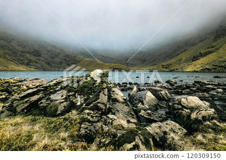 A body of water nestled between a foggy mountain range. The water is calm and reflects the cloudy sky. Rocks and grass are scattered along the shore. 120309150