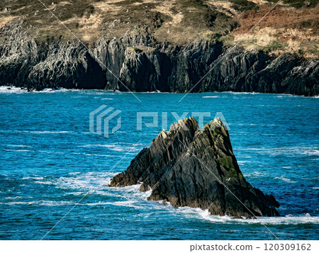 A jagged rocky formation emerges from the clear blue sea, surrounded by gentle waves, with rugged cliffs visible in the background under sunny skies. A jagged rocky formation emerges from the clear blue sea, surrounded by gentle waves, with rugged cliffs visible in the background under sunny skies. 120309162