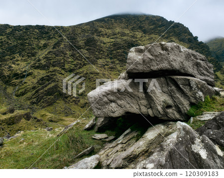 A striking granite boulder rests firmly on a verdant hillside surrounded by rugged terrain. The scene is set against a backdrop of cloudy skies, highlighting the natural beauty. 120309183